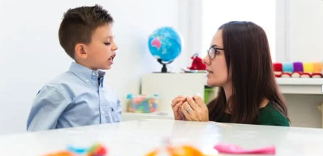 young boy with a blue shirt and black hair is sitting at a table looking at a young woman with glasses and black hair.
